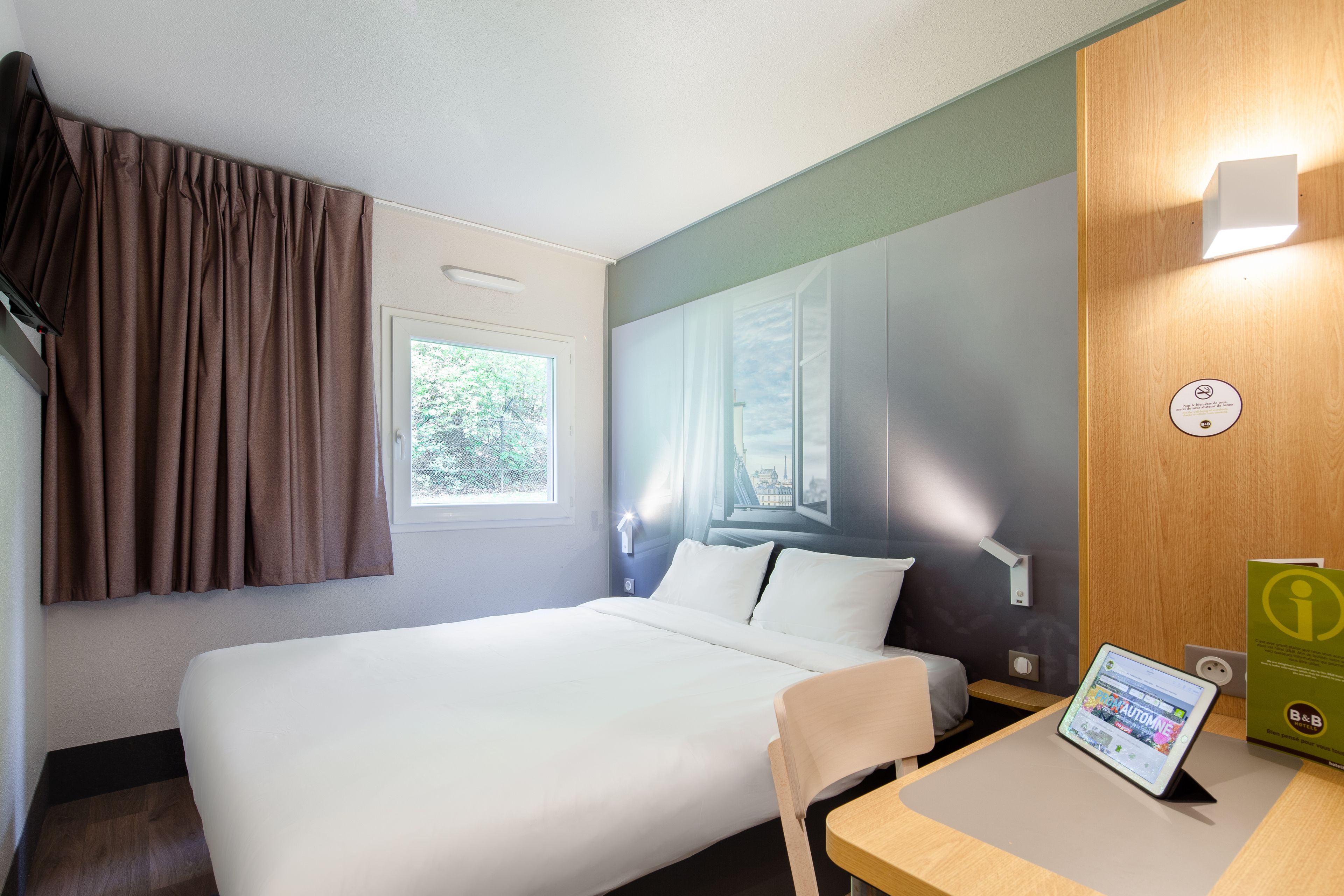 Hotelkamer met een bed, een bureau met een tablet, een raam met gordijnen en een wandlamp.