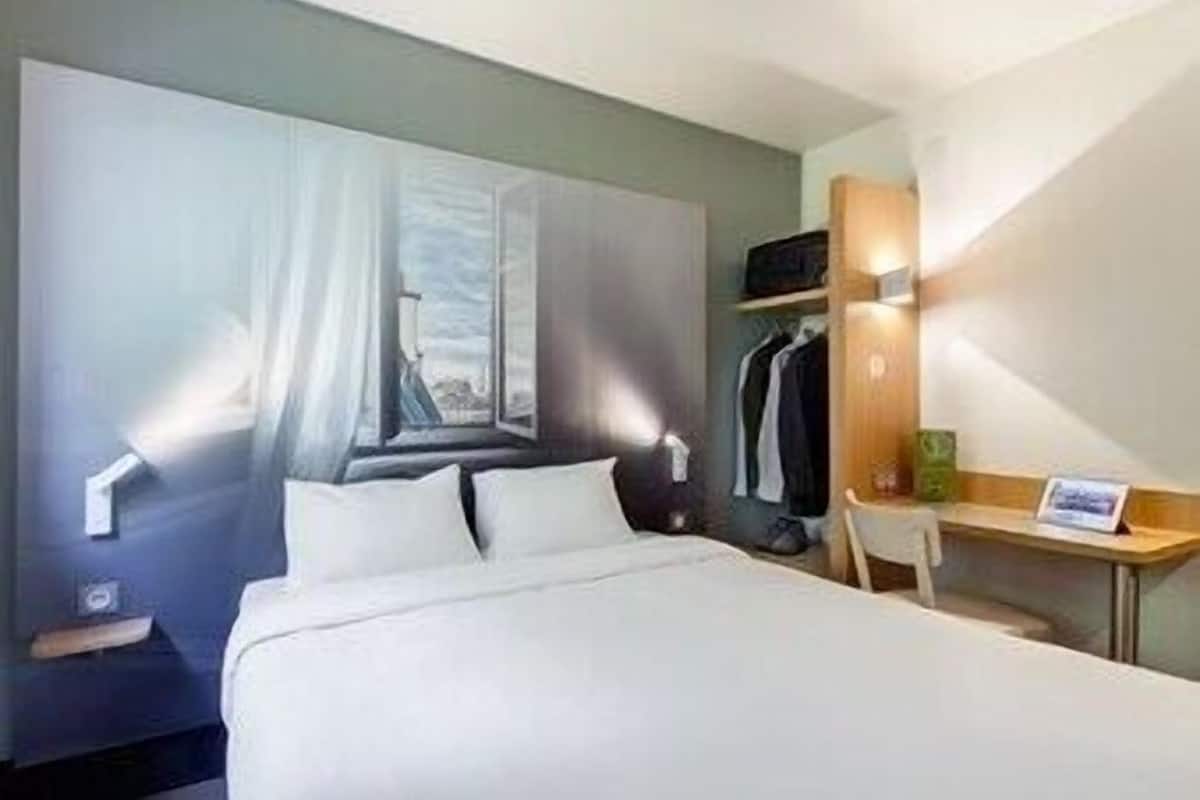 Une chambre d’hôtel équipée d’un lit, d’un bureau, d’une chaise, d’un porte-manteau et d’une grande fenêtre avec des rideaux.