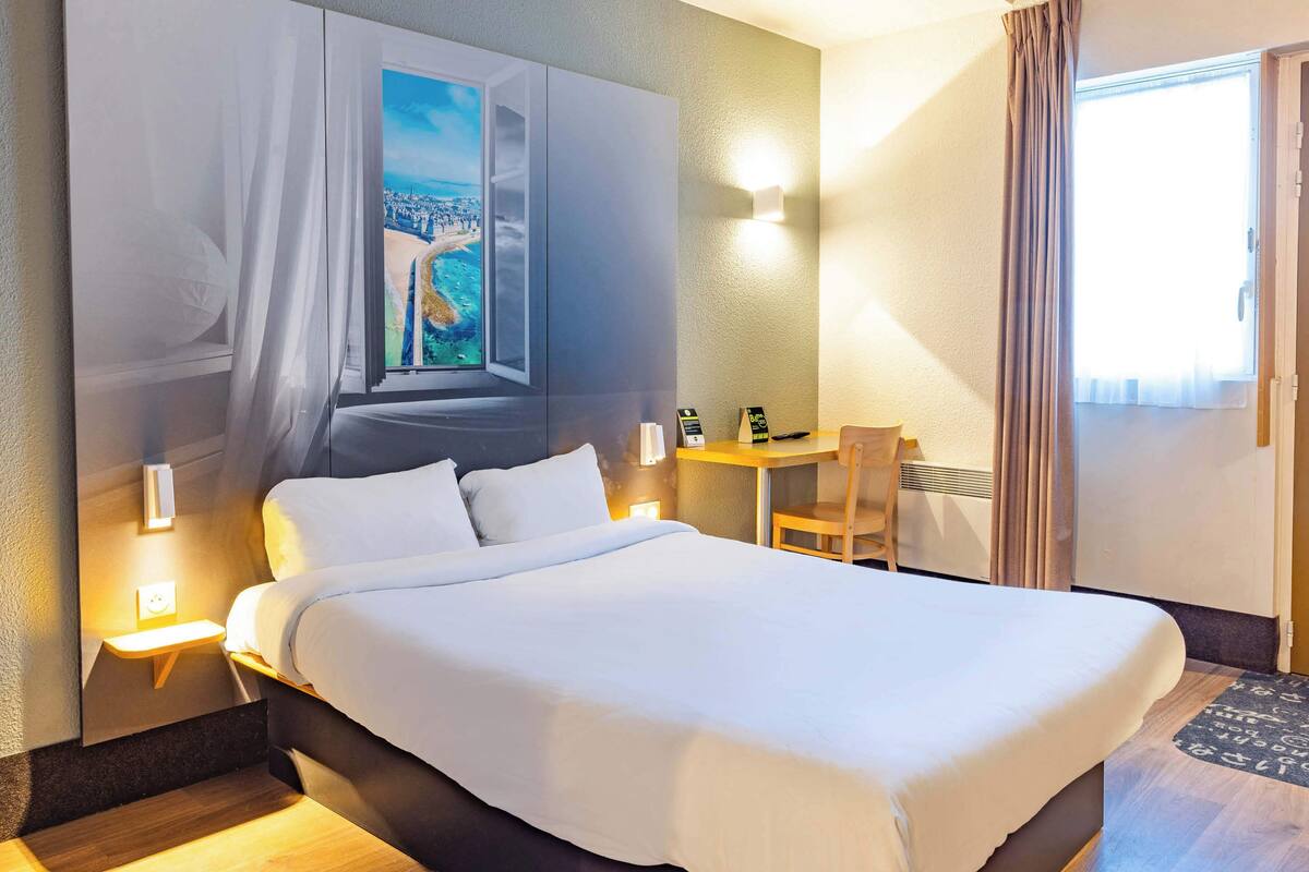 A hotel room with a large bed, a desk, a chair, a window with curtains, and a wall-mounted light.