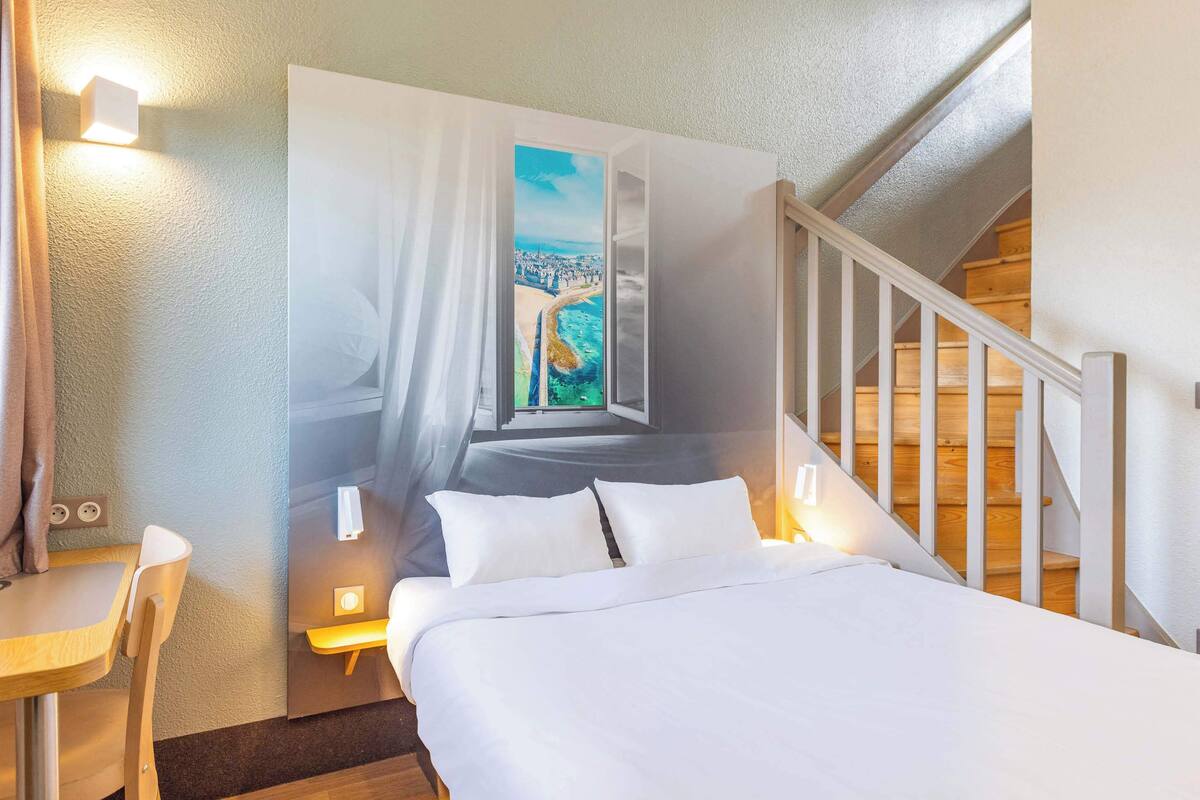 A bedroom with a bed, a desk, a chair, a painting, and a staircase.