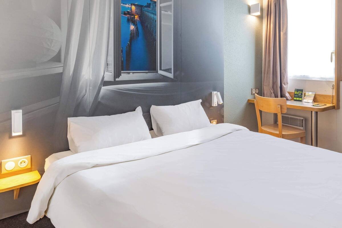 A hotel room with a bed, a nightstand with a lamp, a chair, a desk with a computer, and a window with a view of a bridge.