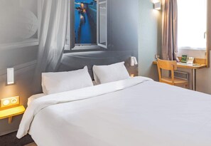 Une chambre d’hôtel comprenant un lit, une table de chevet avec une lampe, une chaise, un bureau avec un ordinateur et une fenêtre donnant sur un pont.