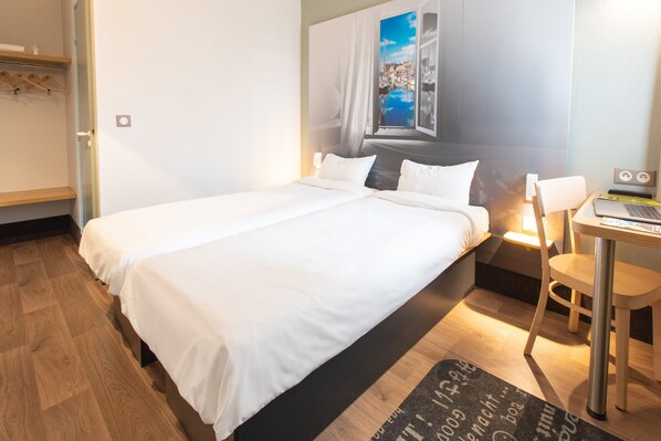 A hotel room with a bed, a desk with a laptop, a chair, a window with a view of a cityscape, and a wooden floor.