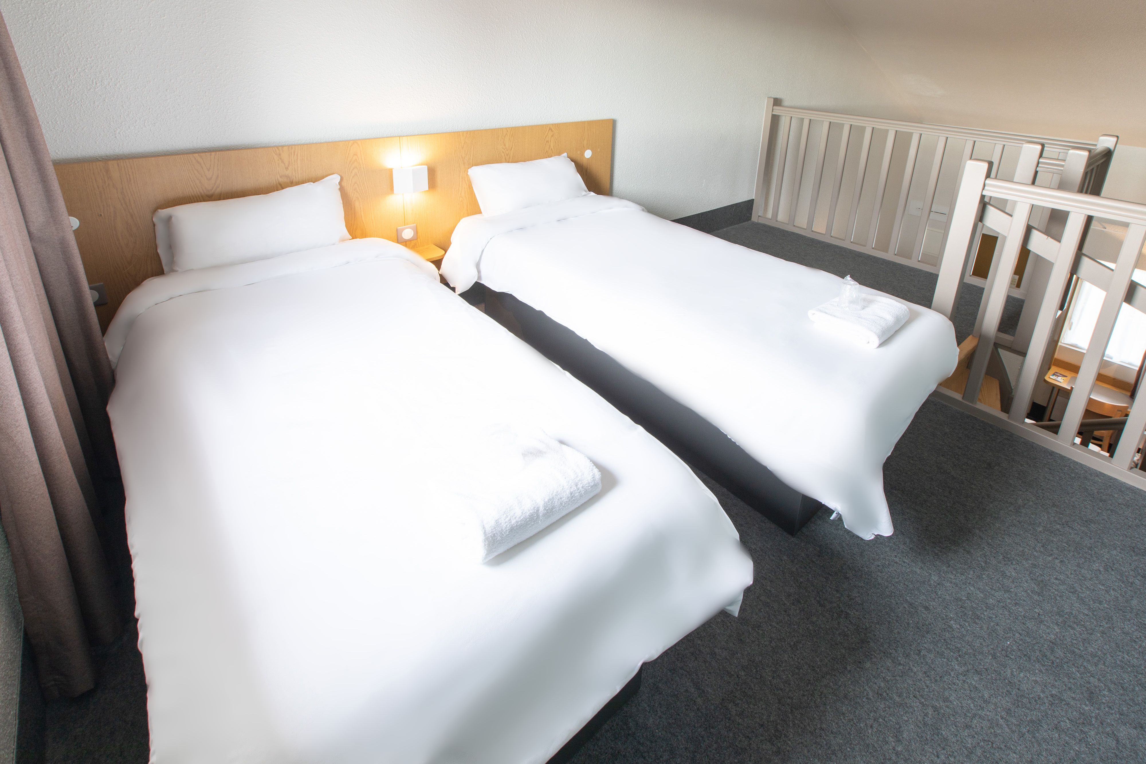 Habitación de hotel con dos camas individuales, cada una con cubrecama y almohadas blancas, cabecera de madera y mesita de noche con lámpara.