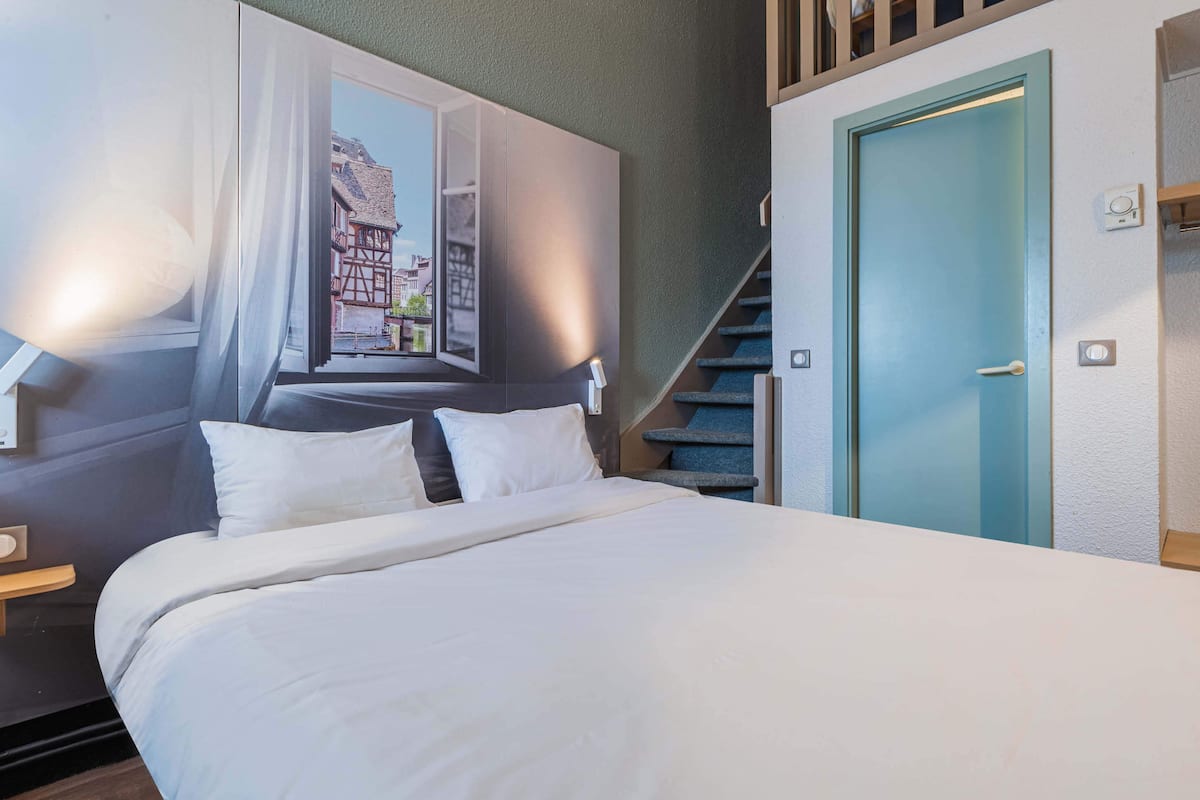 Habitación de hotel con cama, ventana con cortinas, vista a edificios, una puerta azul y una escalera.