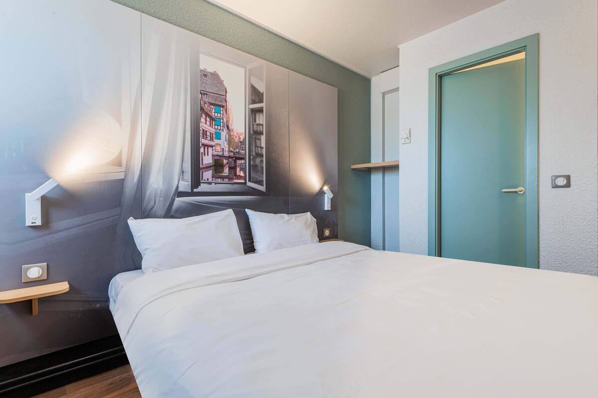 Una habitación de hotel con cama, ventana con cortinas, puerta y mesita de noche.
