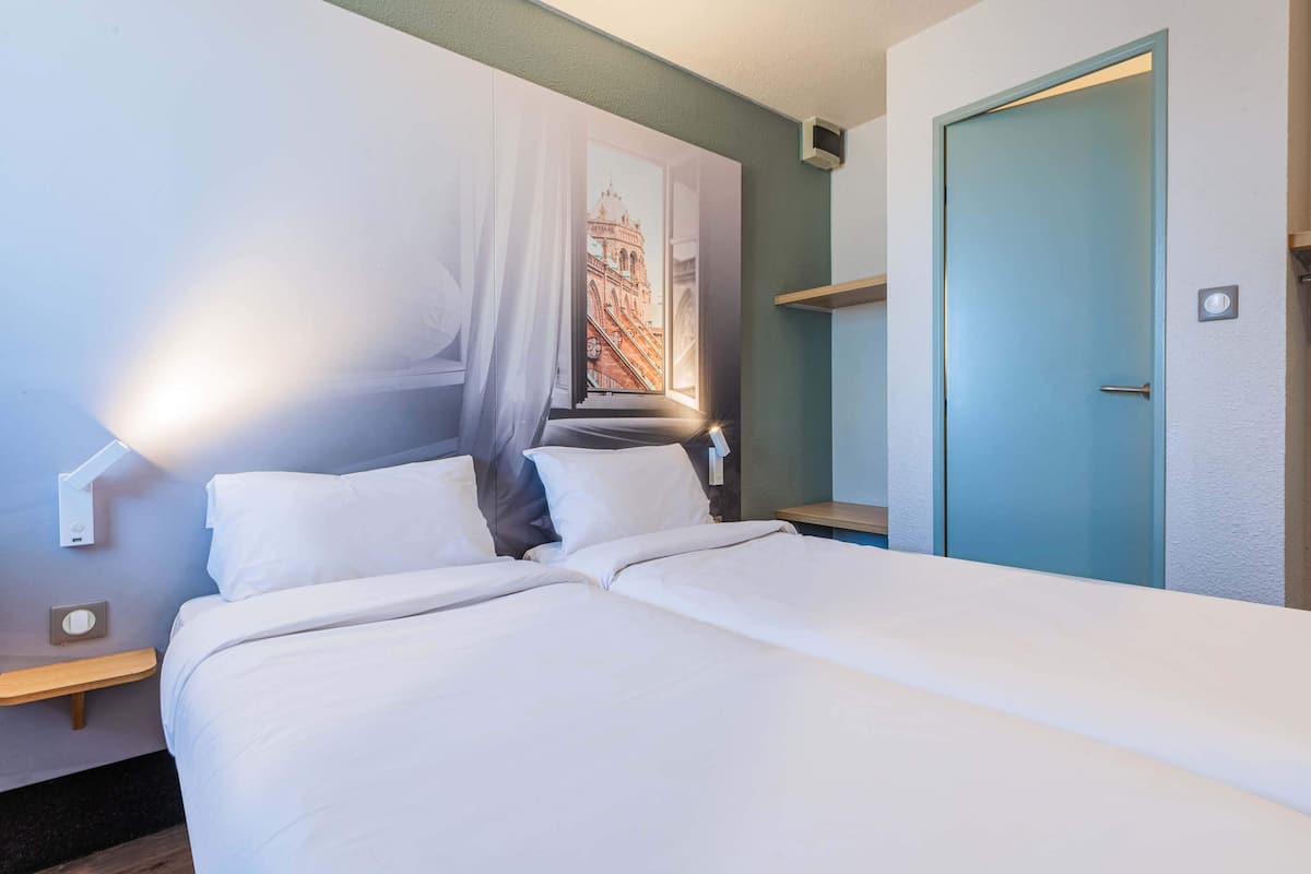 Una habitación de hotel con una cama, una mesita de noche, una lámpara de pared, una puerta y una ventana con vista a la ciudad.