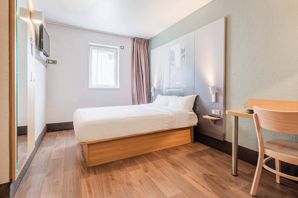 Un dormitorio con una cama, una mesita, una silla, un televisor en la pared y una ventana con cortinas.