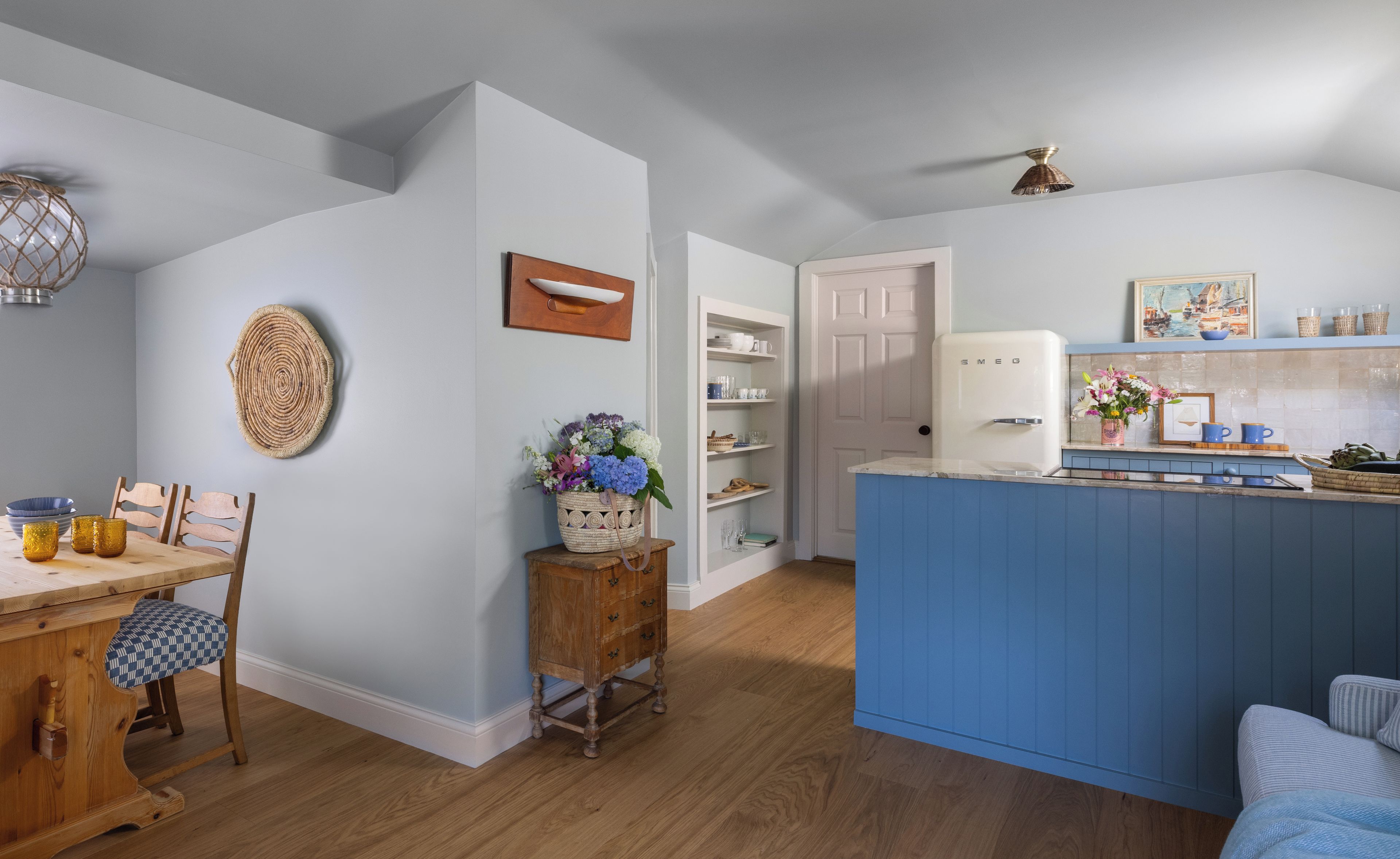 A modern kitchen with a blue island, wooden chairs, a dining table, and a white refrigerator.