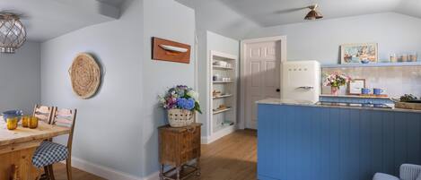 A modern kitchen with a blue island, wooden chairs, a dining table, and a white refrigerator.