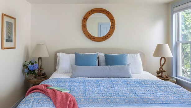 A bedroom with a bed, two bedside tables, a round mirror, a window, and a framed picture on the wall.