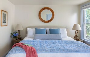 A bedroom with a large bed, two bedside lamps, a round mirror, and a window with a view of greenery. - The Lightkeepers Inn (Edgartown)
