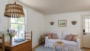 A room with a wooden desk, a white sofa with patterned pillows, a wicker lamp, and a framed picture on the wall.