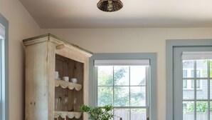 A room with a wooden cabinet, a table with chairs, a hanging light fixture, and a window with a view of greenery.