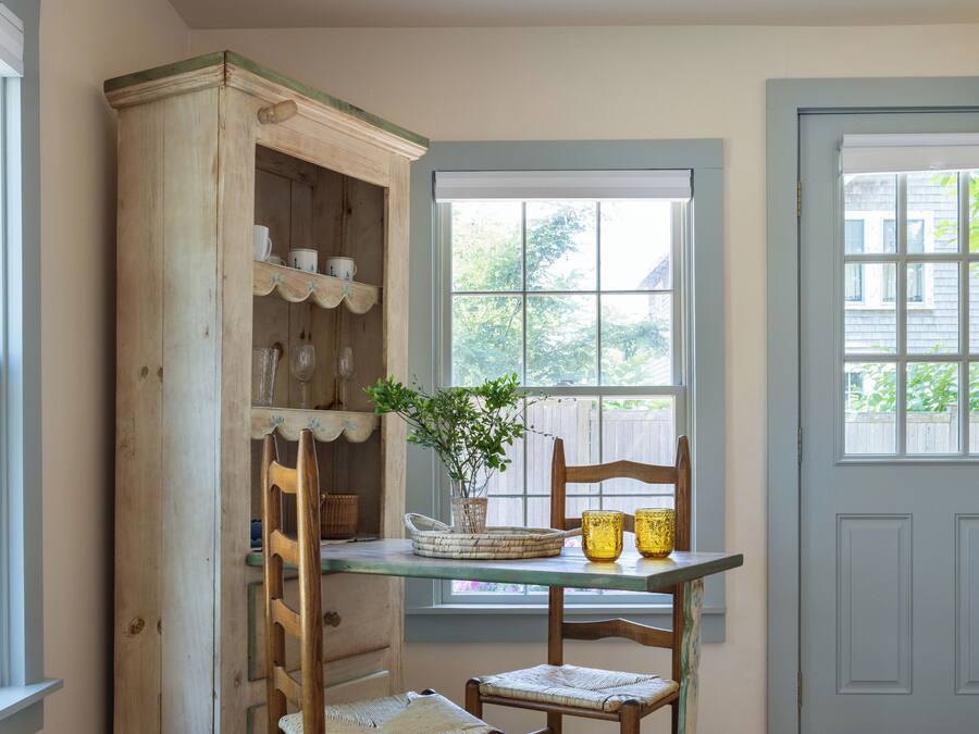 A room with a wooden cabinet, a table with chairs, a hanging light fixture, and a window with a view of greenery.