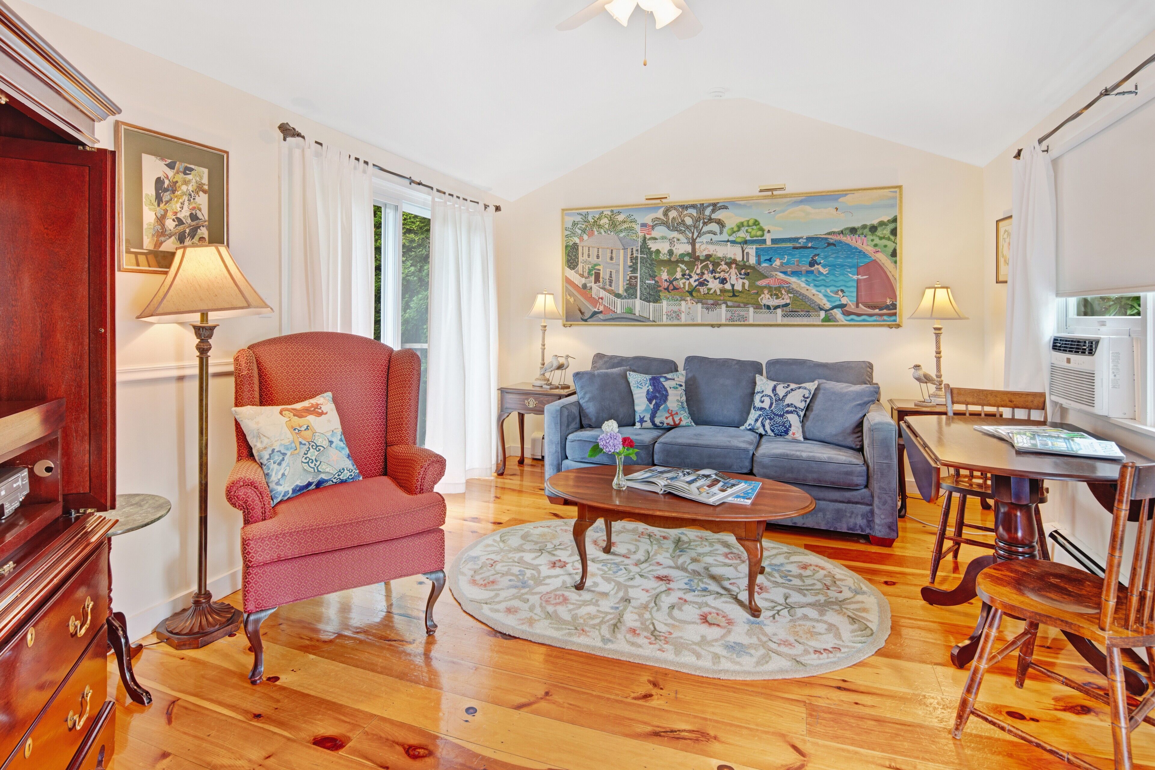 A living room with a red armchair, a blue sofa, a coffee table, and a painting of a coastal scene.