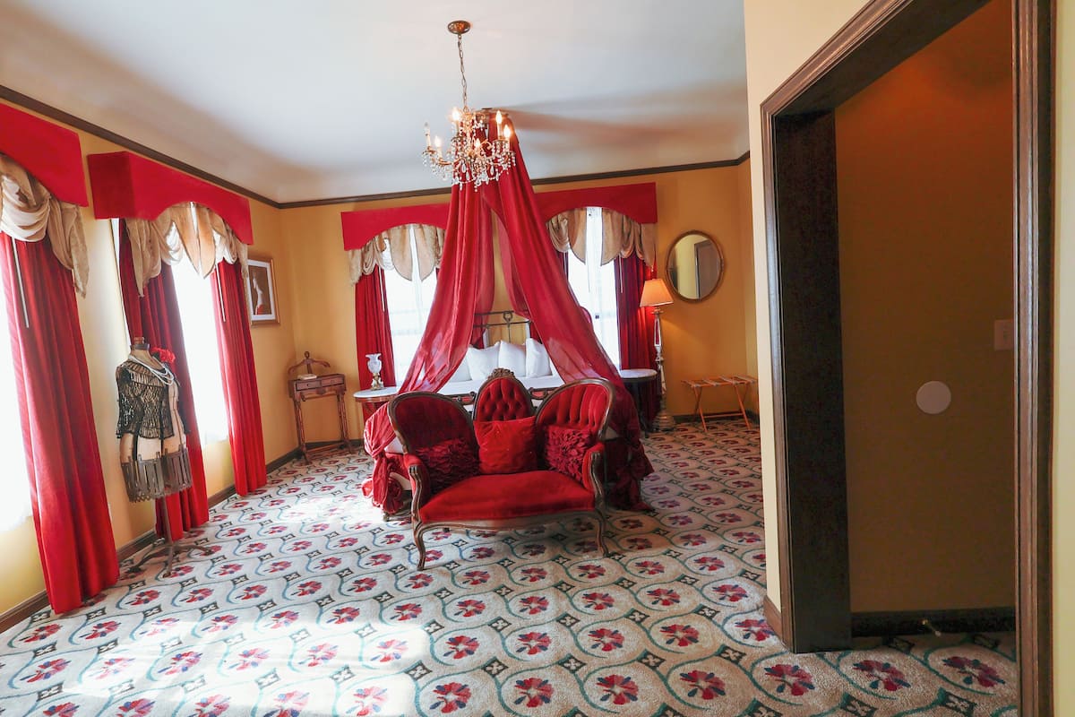 A room with a canopy bed, red curtains, a chandelier, and patterned carpet.