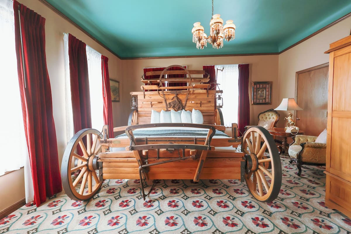A room with a wooden bed featuring a large wooden wheel, red curtains, a chandelier, and a patterned carpet.