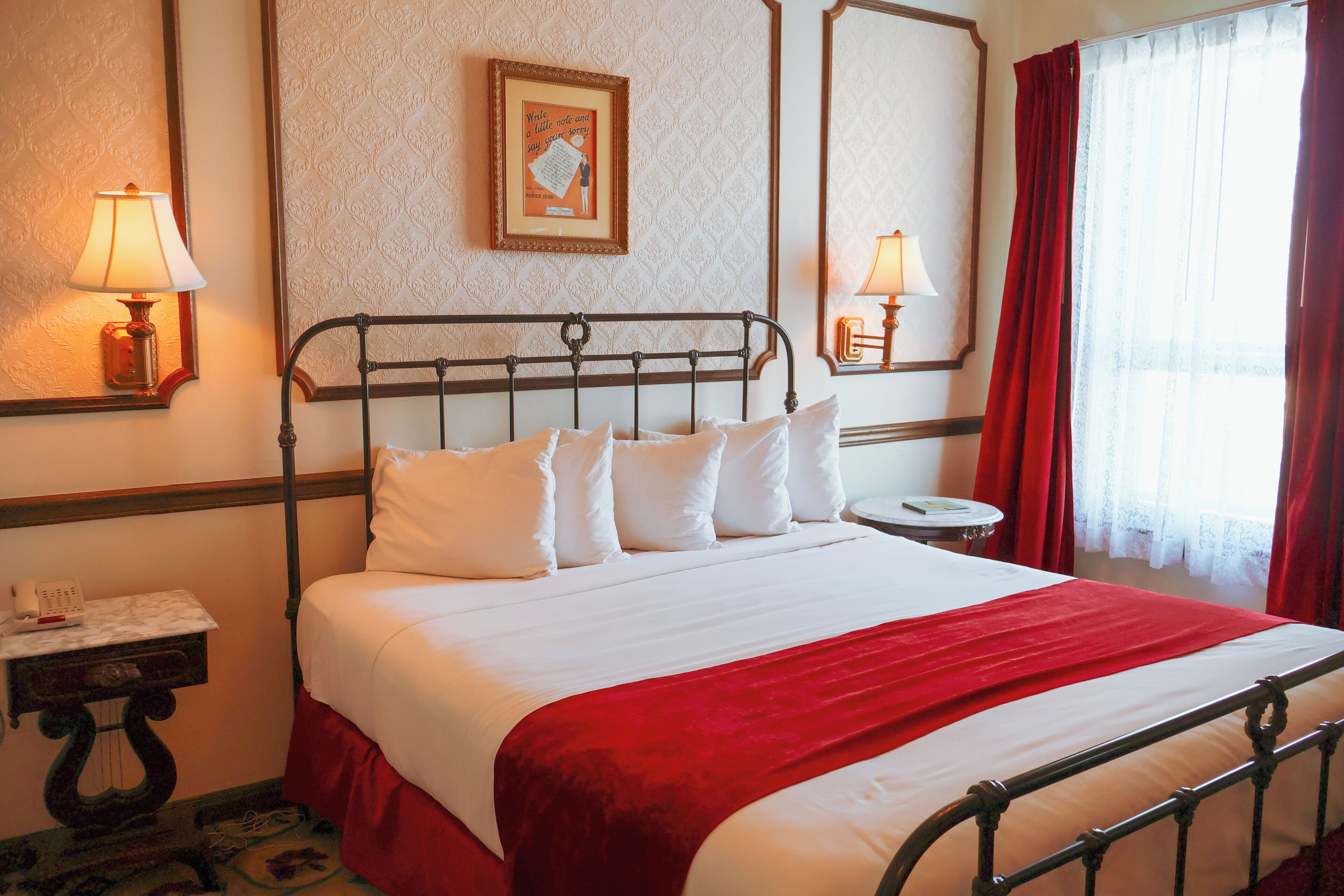 A neatly made bed with white linens and a red blanket, a bedside table with a lamp, and a framed picture on the wall.