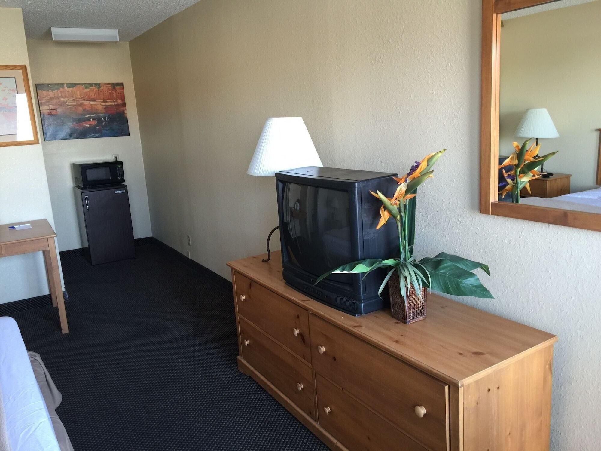 A hotel room with a wooden dresser, a television, a lamp, a small refrigerator, and a painting on the wall.