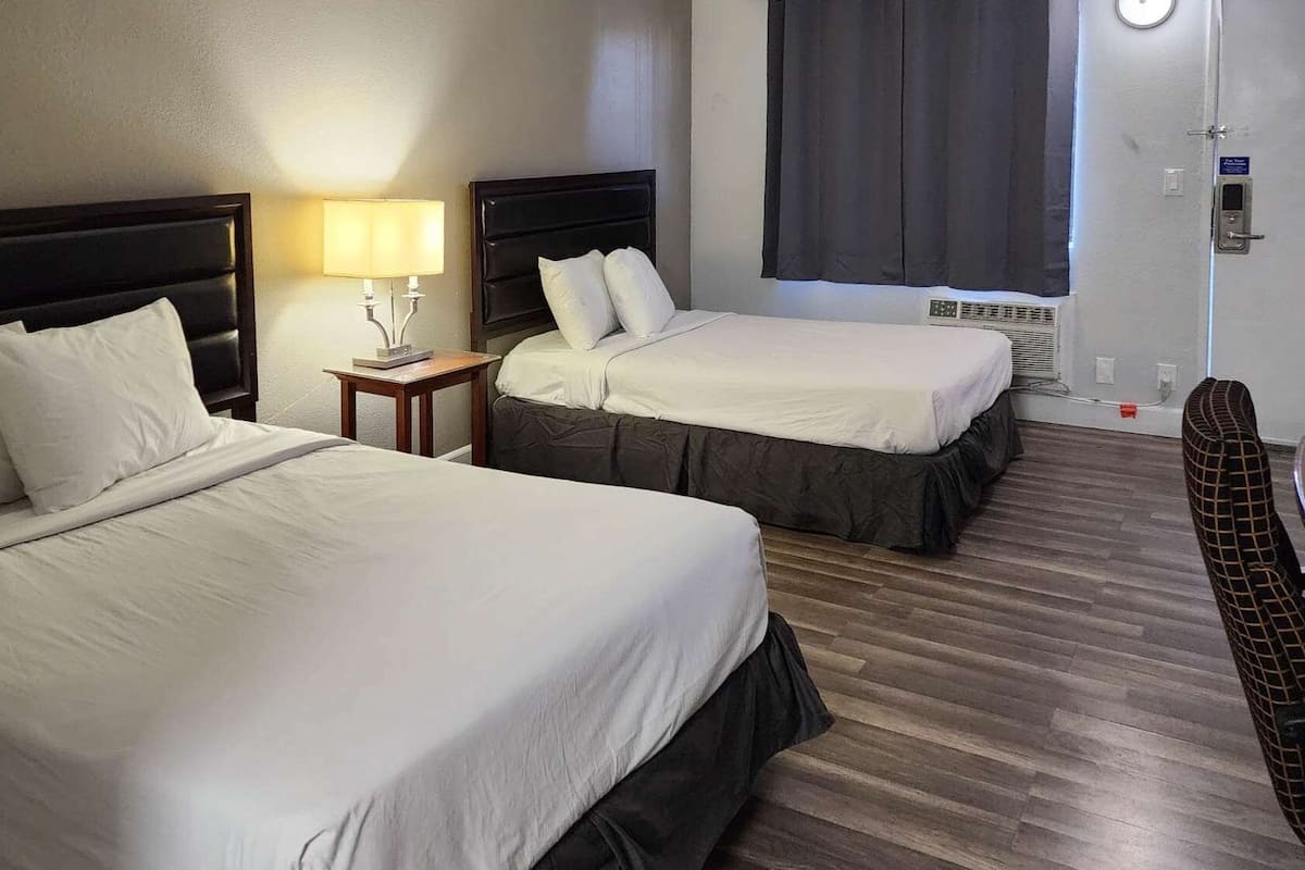 A hotel room with two beds, a nightstand with a lamp, a window with curtains, and a door with a lock.