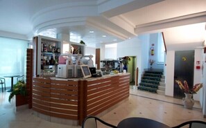 Zona de recepción con mostrador de madera, máquina de café y exhibición de bebidas embotelladas. Se ve una escalera que sube a otro nivel y una pequeña mesa con sillas en primer plano.