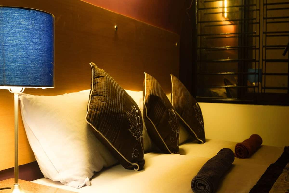 Ein ordentlich bezogenes Bett mit einem weißen Kopfkissen, drei dekorative Kissen mit Blättermotiven, ein aufgerolltes Handtuch und eine blaue Lampe auf einem Nachttisch.
