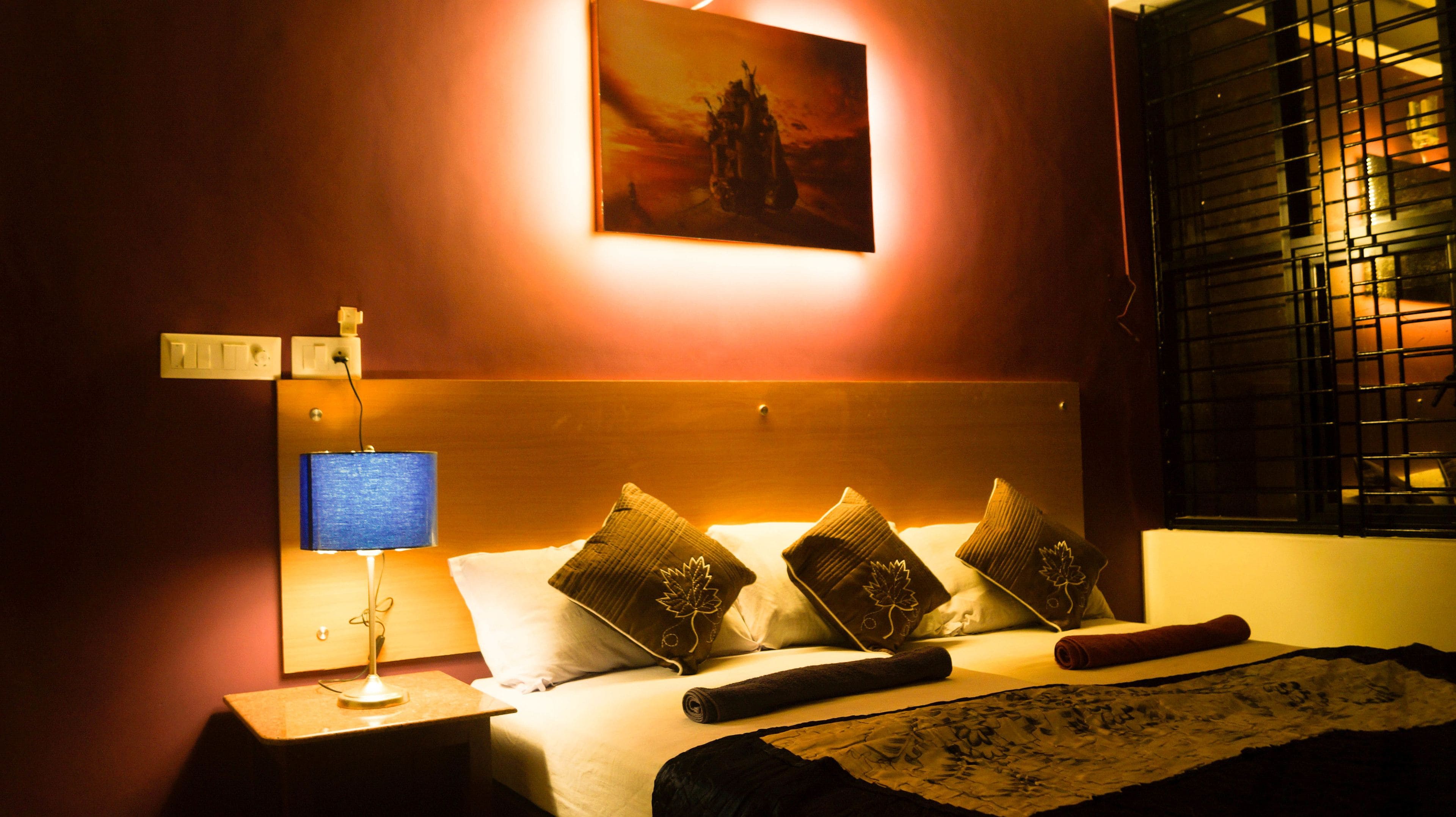 Een hotelkamer met een bed, kussens, een nachtkastje met een lamp, een schilderij aan de muur en een raam met jaloezieën.