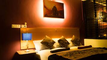Quarto de hotel com cama, almofadas, mesa de cabeceira com abajur, quadro na parede e janela com persianas.