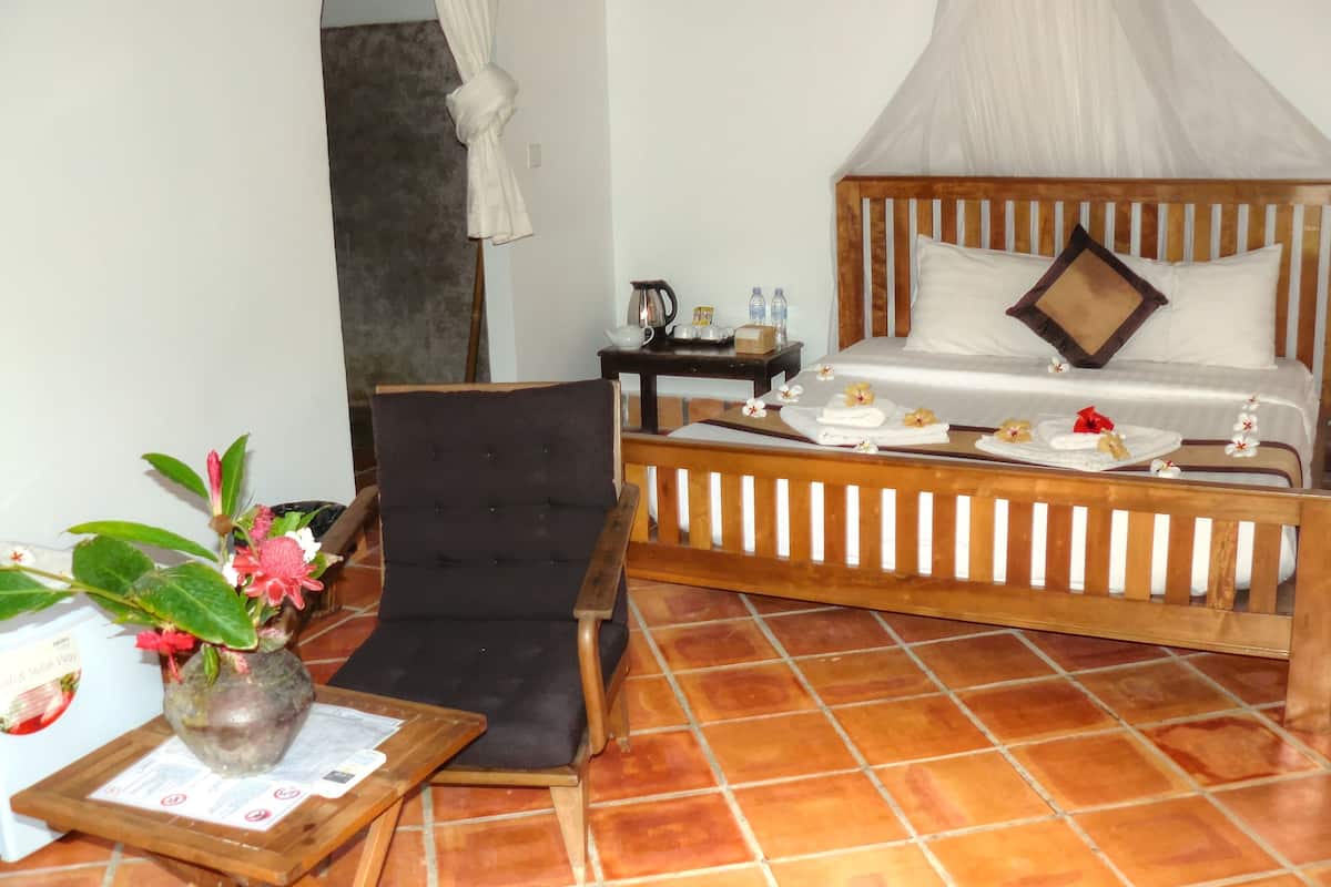 Habitación de hotel con cama, una silla, una mesita con un florero y piso de baldosas.