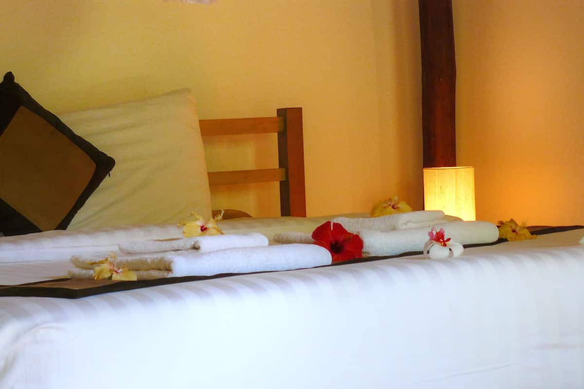Una cama bien tendida con toallas y flores, cabecera de madera y lámpara de noche.