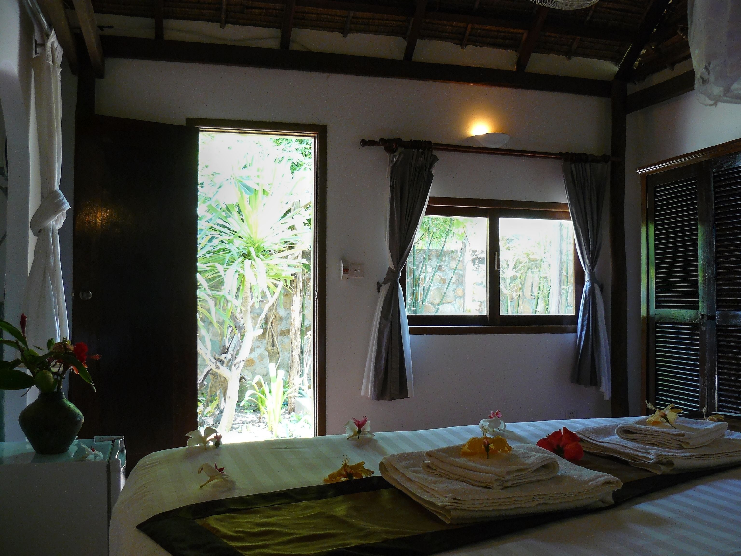 A hotel room with a bed, a window with curtains, a bedside table with a vase of flowers, and a view of greenery outside.