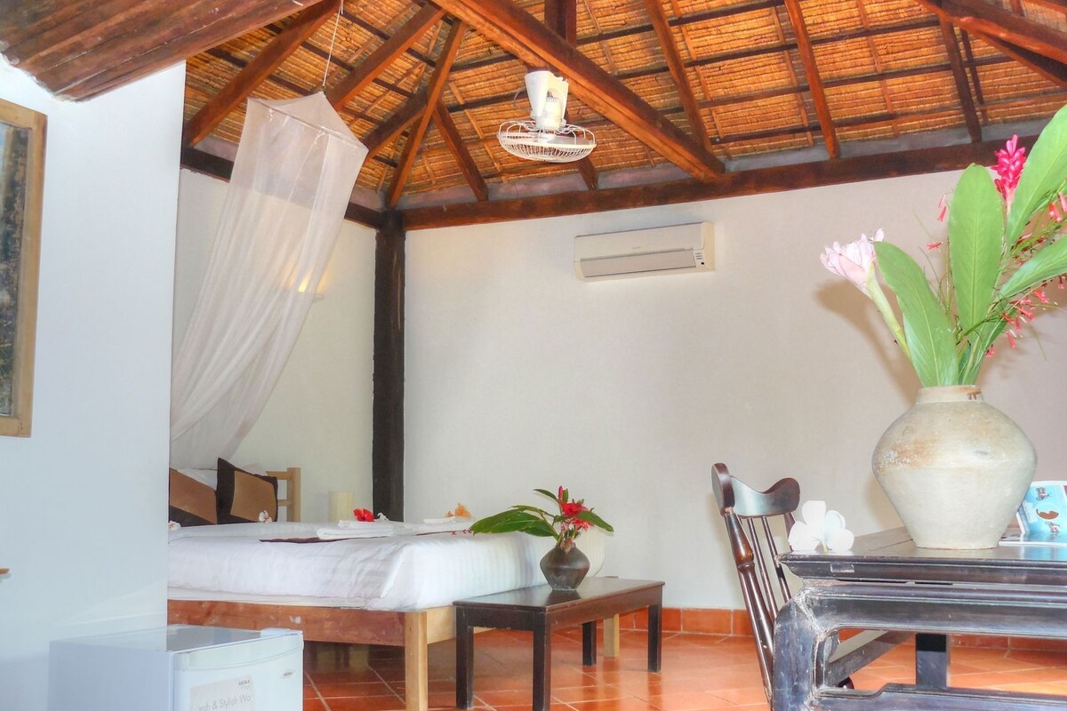 Quarto com cama, mesa, cadeira, vaso com flores e ventilador de teto.