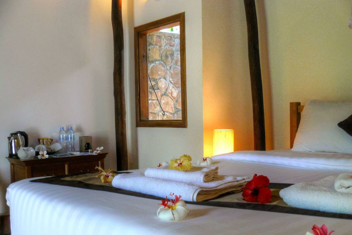 Habitación de hotel con cama, mesita de noche, lámpara y ventana con vista a un muro de piedra.