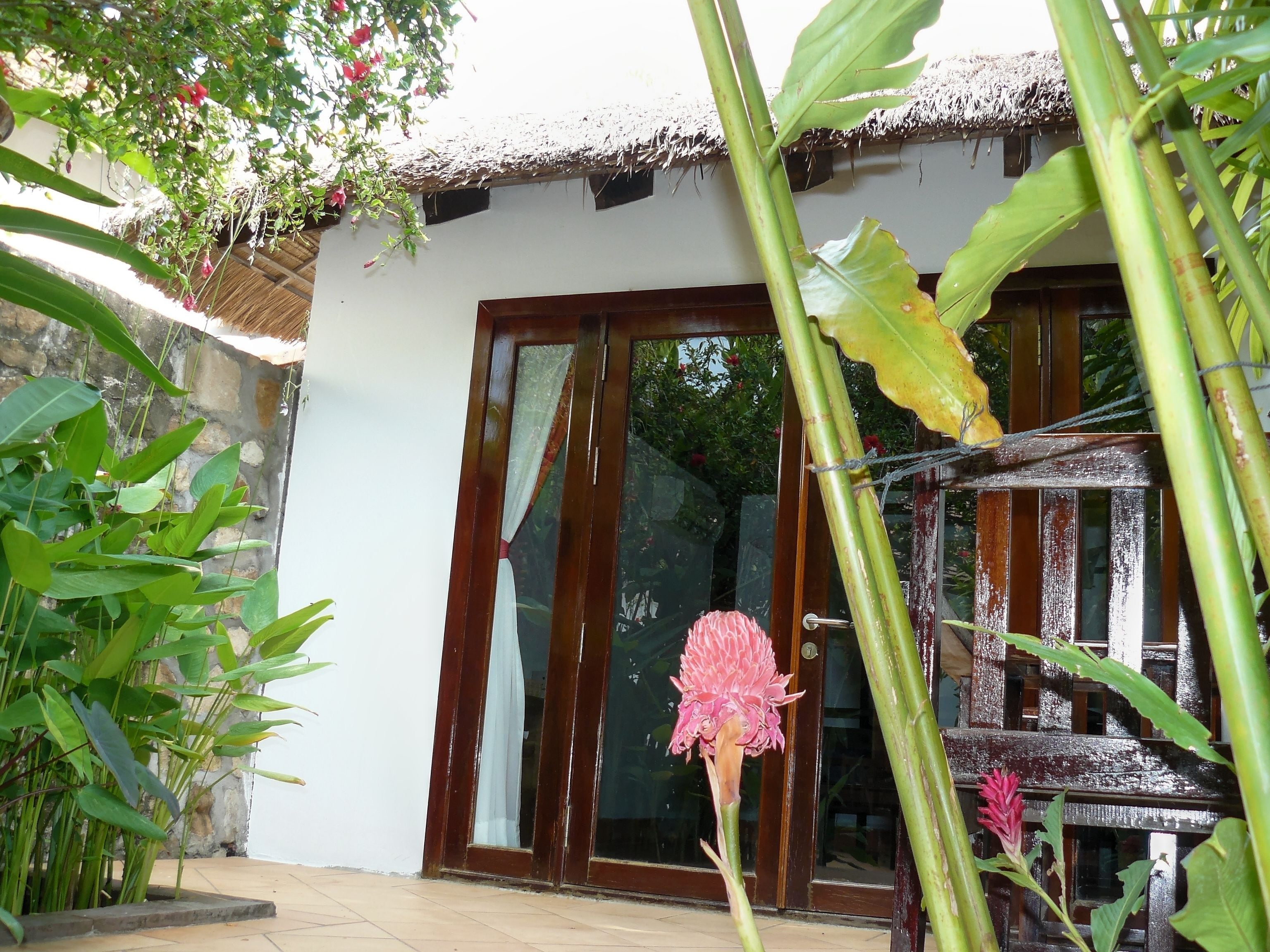 Uma casa com telhado de palha, grandes portas de vidro e um jardim com várias plantas e uma flor rosa.