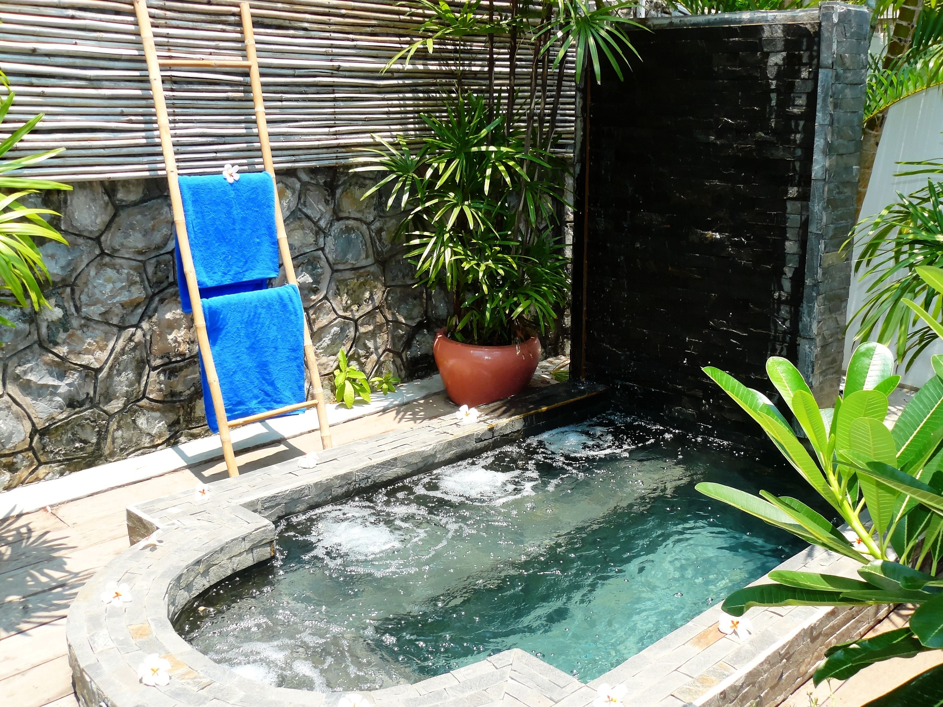 Un área con jacuzzi de paredes de piedra, una escalera de bambú, una planta en maceta y una toalla azul colgada en la pared.