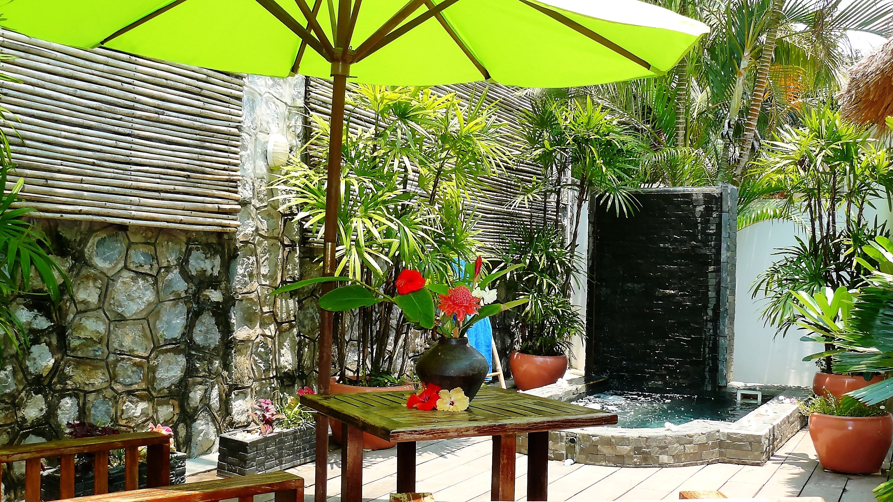 A patio area with a stone wall, a wooden table with a vase of flowers, a green umbrella, and a small water feature.