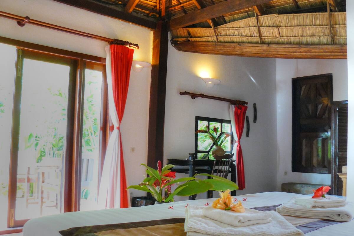 Una habitación con cama, ventana con cortinas rojas, una planta y una mesa con toallas.