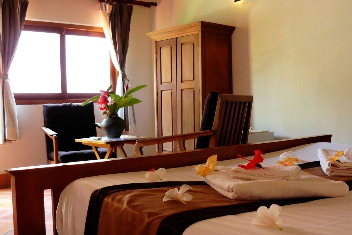 Habitación de hotel con cama, una silla, una mesita con un florero y cortinas, un armario y una ventana.