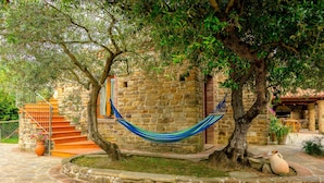 Un bâtiment en pierre avec un hamac suspendu entre deux arbres, un escalier et un espace patio agrémenté de plantes en pot.