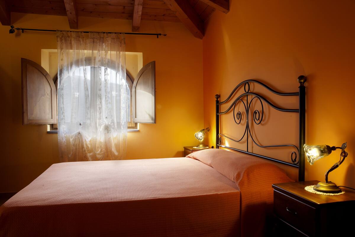 Ein Schlafzimmer mit einem Bett, einem Nachttisch, einer Lampe und einem Fenster mit Vorhängen.