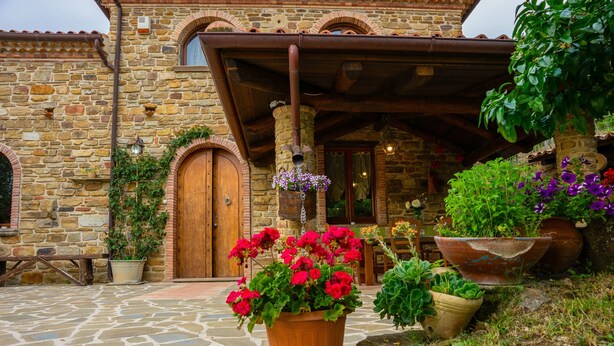 Un bâtiment en pierre avec une porte en bois et un porche. Des plantes en pot et des fleurs sont disposées devant le bâtiment.