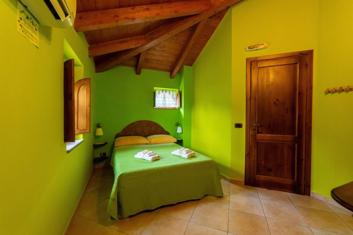 Ein Zimmer mit einem Bett, Holzbalkendecke, grüner Wand, Fenster und Tür.