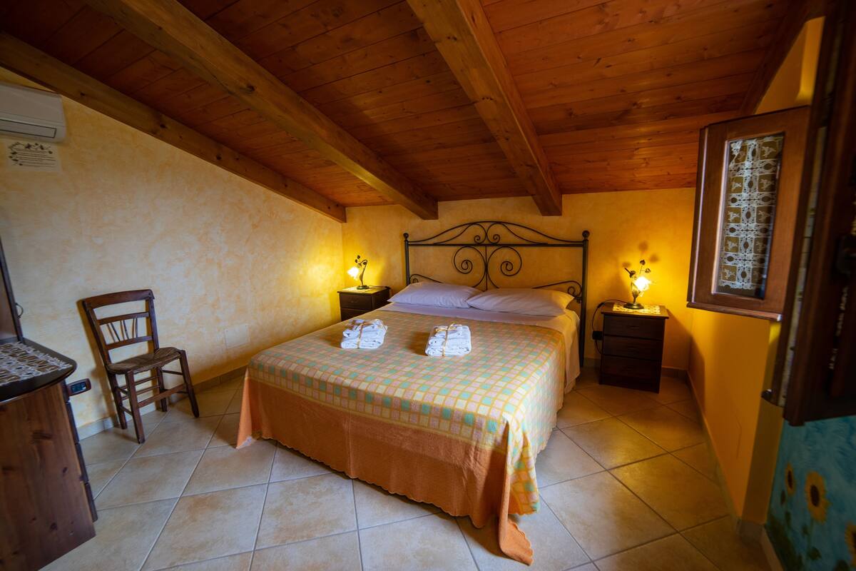 Ein Schlafzimmer mit Holzbalkendecke, einem Bett mit kariertem Überwurf, zwei Nachttischlampen, einem Sessel und einem Nachttisch mit Lampe.