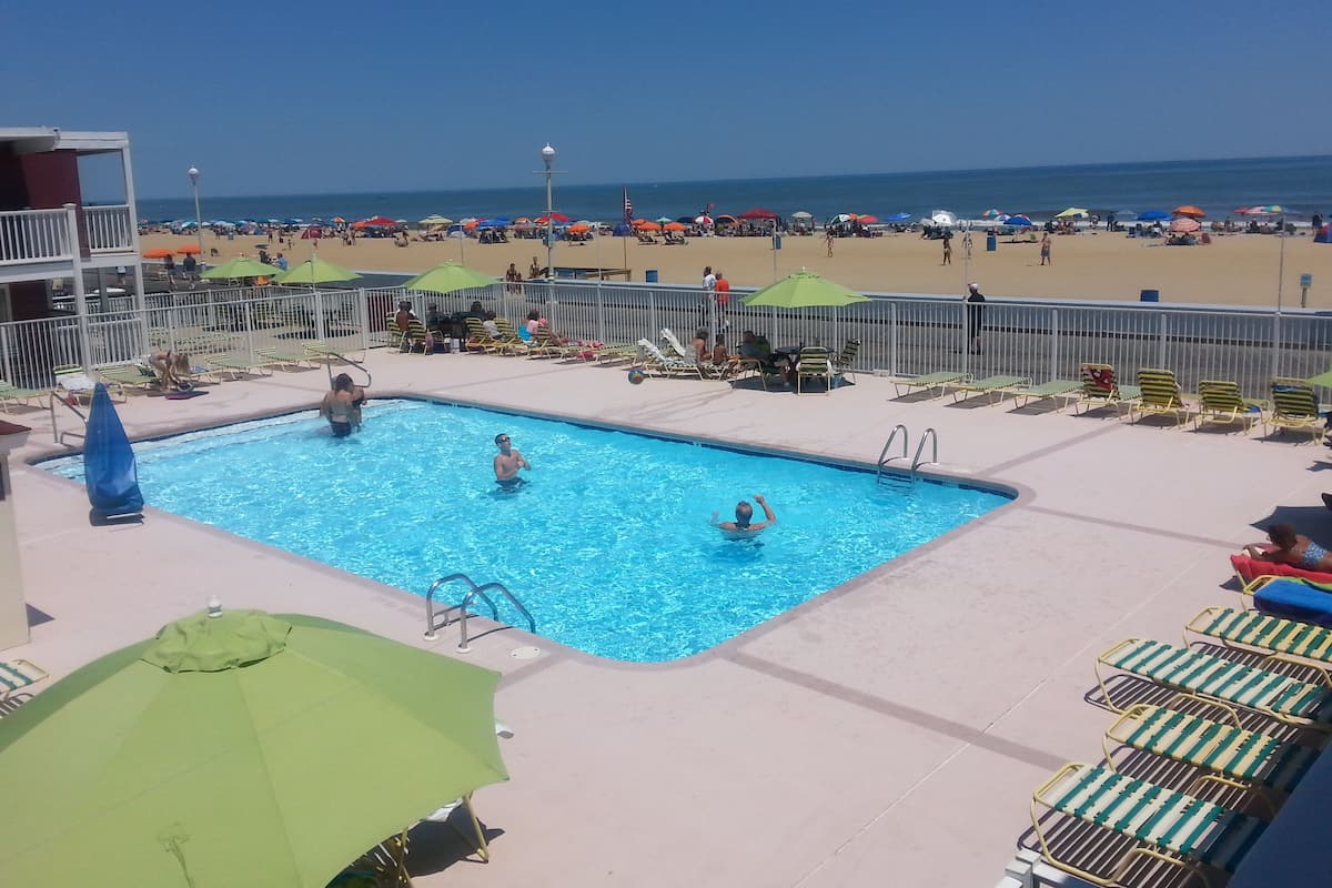 A swimming pool with lounge chairs and umbrellas, a beach in the background, and a clear sky.