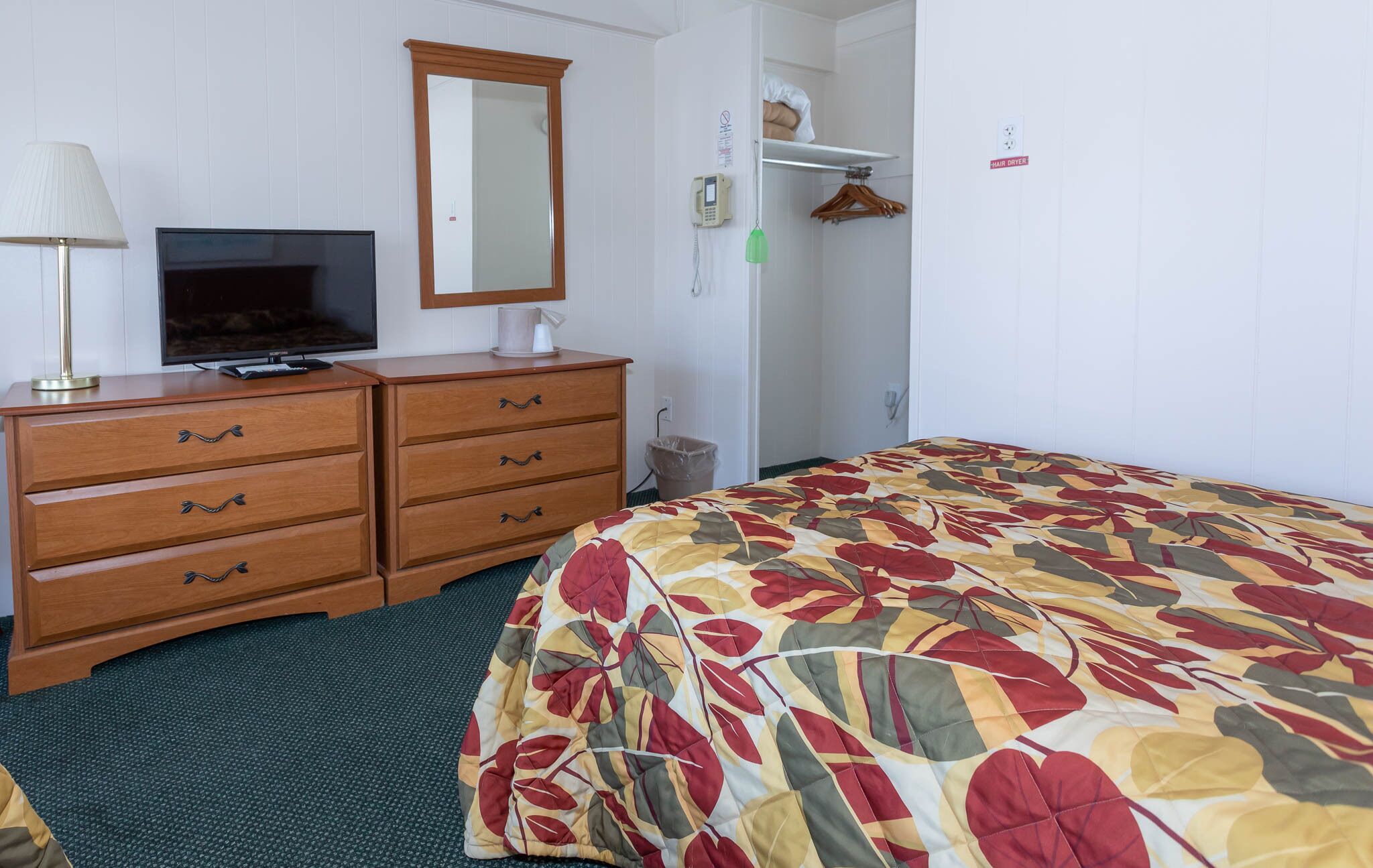 A hotel room with a bed, a dresser with drawers, a television, a mirror, and a hair dryer sign.