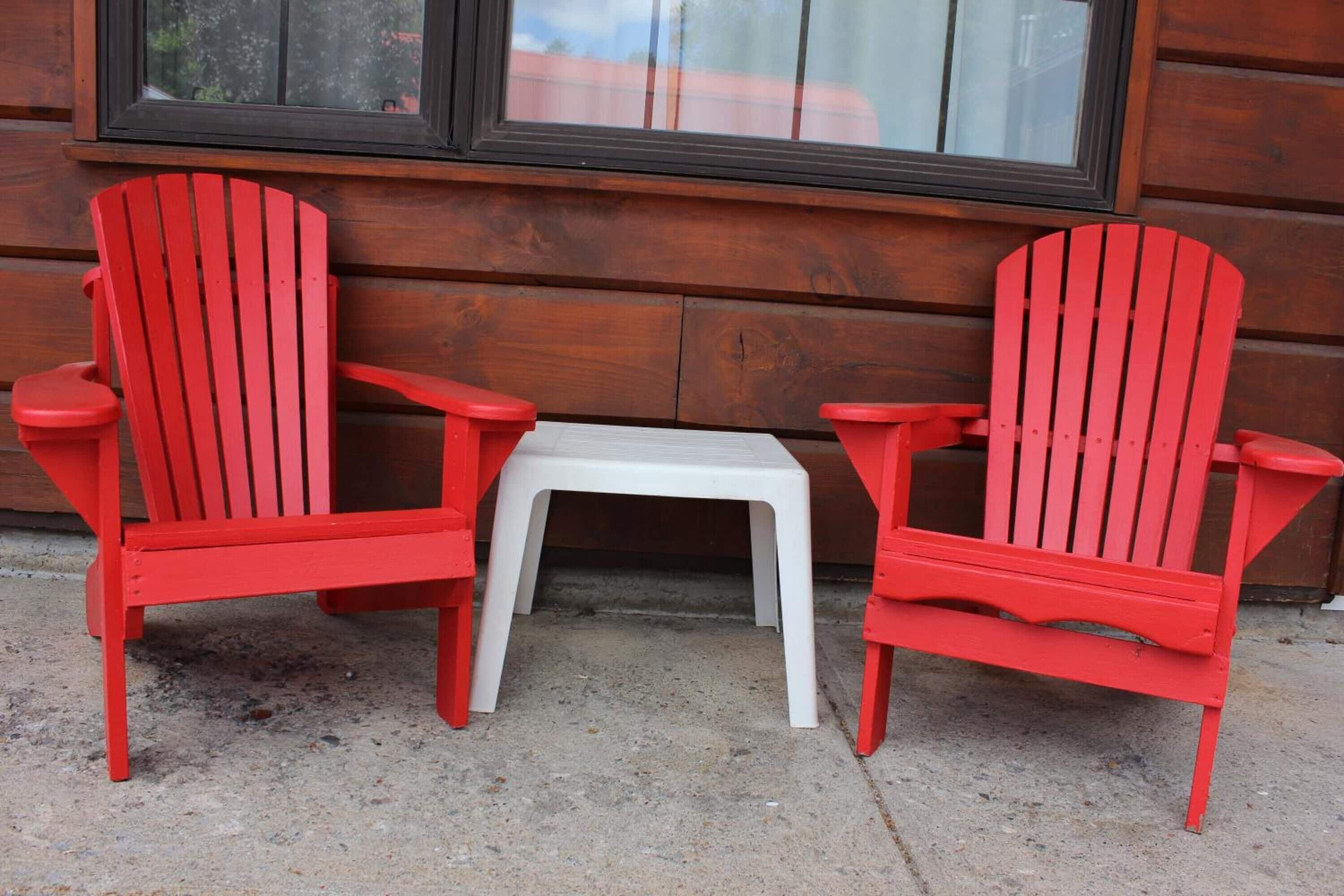 Two red Adirondack chairs and a small white table set against a wooden wall with a window above.