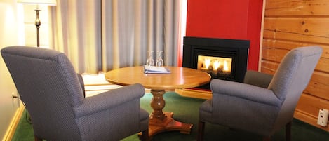 A cozy room with a round wooden table, two patterned armchairs, a fireplace, and a lamp.