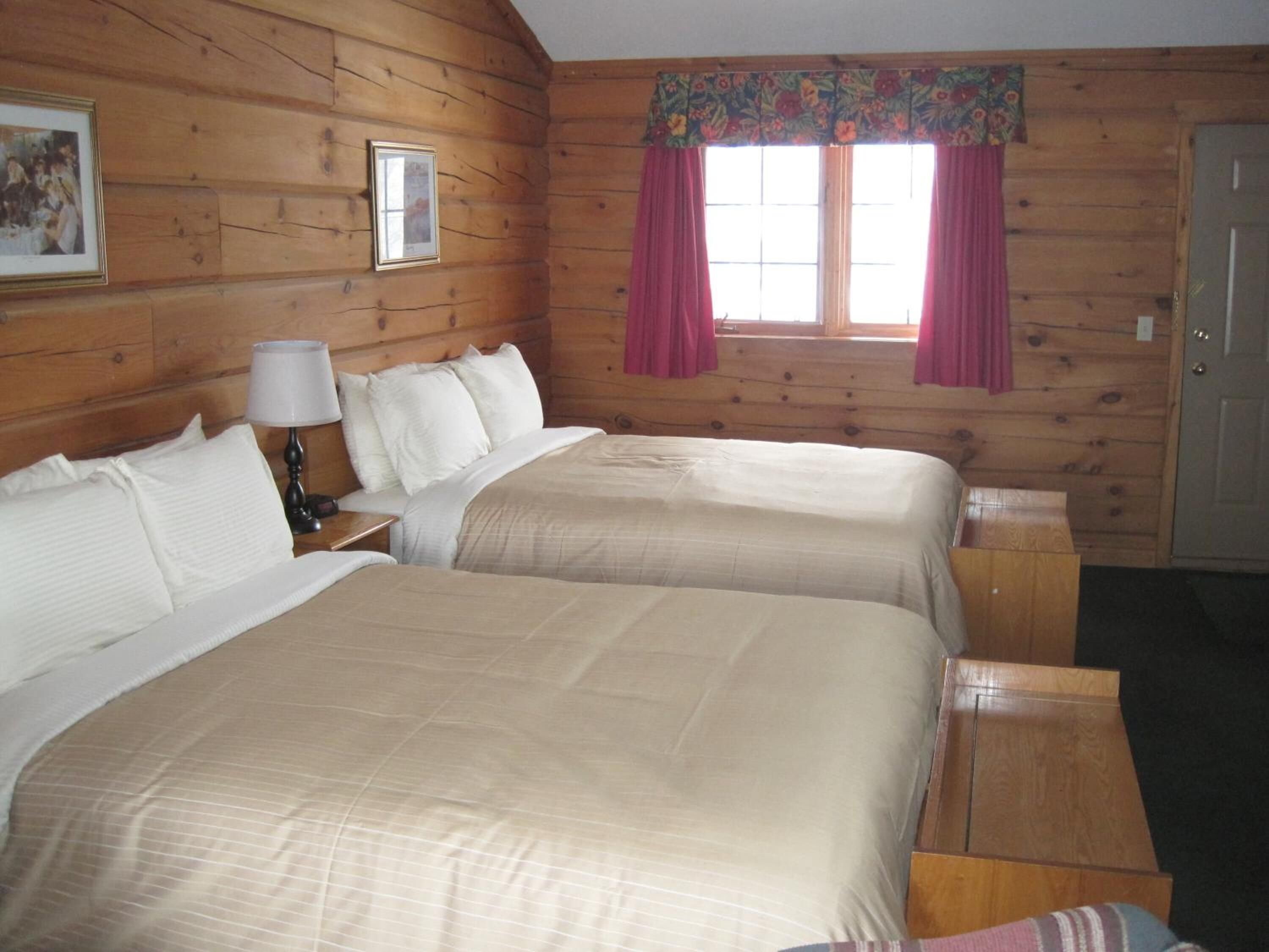 A wooden cabin room with two beds, a nightstand, a lamp, a framed picture, and a window with curtains.