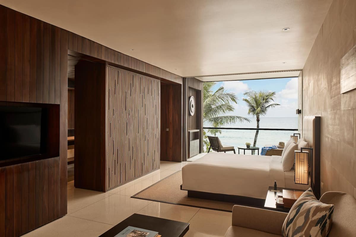 A modern hotel room with a large bed, a TV, a coffee table with books, and a balcony with a view of the ocean.